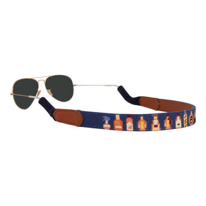Bourbon bottle themed needlepoint sunglass strap showing popular bourbons against a navy blue background with sturdy cotton covered silicone ear connectors and a soft leather backing