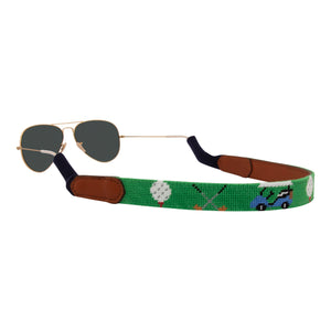 golfing theme handmade needlepoint sunglass strap showing golf carts golf clubs golf balls against a green background with sturdy cotton covered silicone ear connectors and a soft leather backing