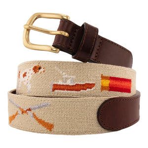 Sportsman Needlepoint Belt with khaki colored background showing outdoor sporting themed images of a pointer dog, crossed shotguns, shotgun shell, fly fishing rod, redfish, fishing boat, flying duck and dove.  Belt has brass buckle and full-grain leather backing.