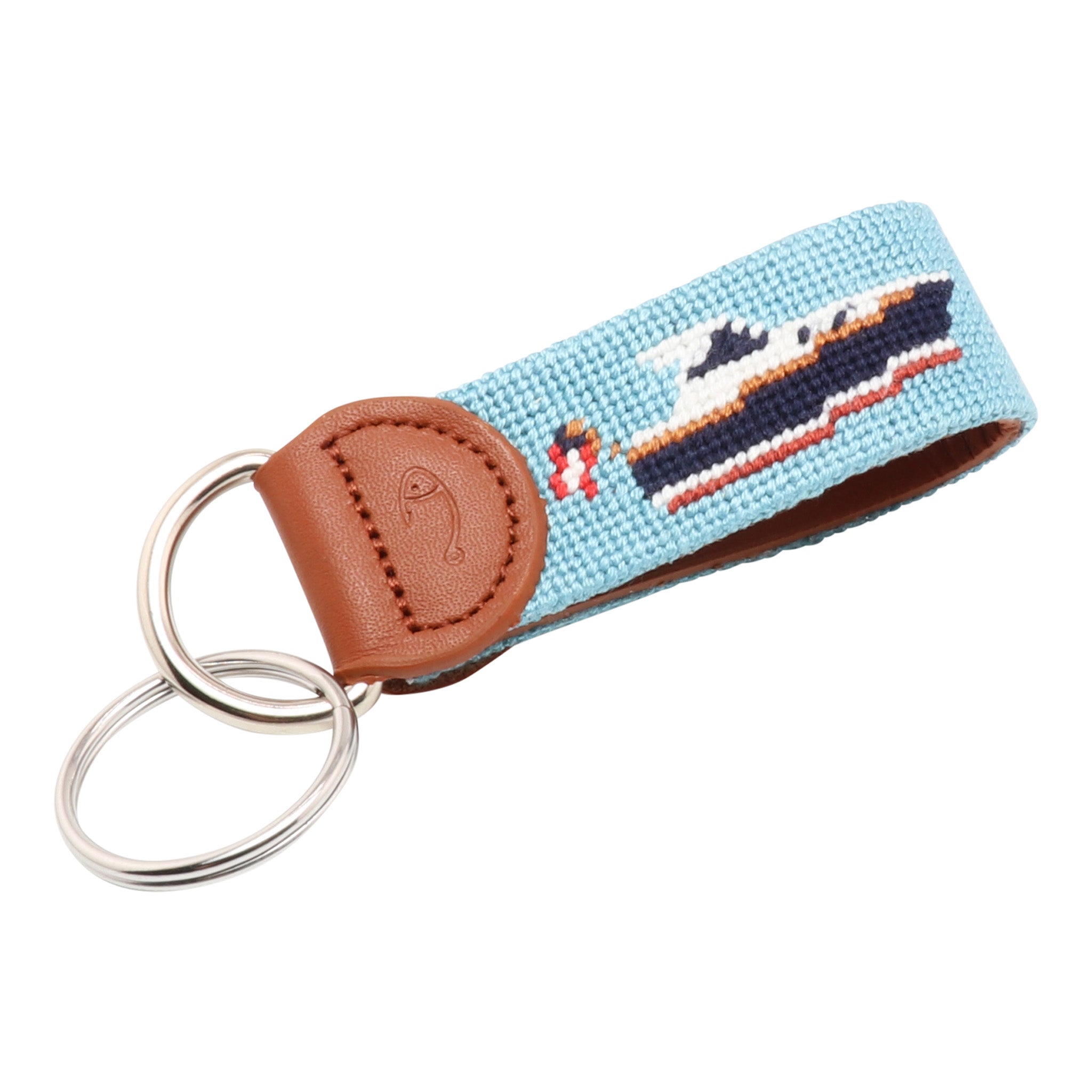 A handcrafted key fob featuring a needlepoint design with a boat on a blue background, attached to a stainless steel keyring and a leather strap.