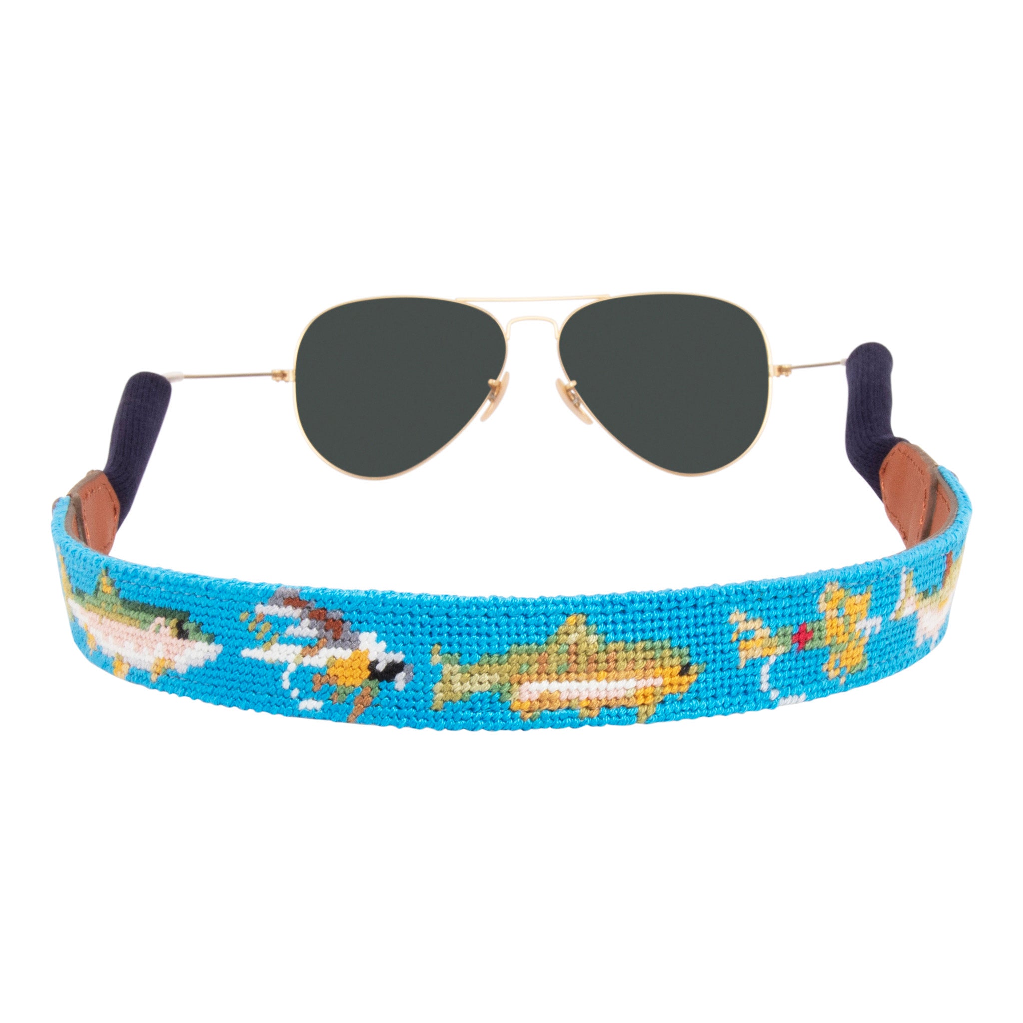 Fly Fishing Needlepoint Sunglass Strap with light blue background showing a fly-fishing themed pattern of rainbow trout, salmon, fly rods and reels, dry flies, nymph patterns, and cutthroat.  Sunglass straps have a full grain leather backing and secure cotton wrapped ear piece connectors for a secure grip. 
