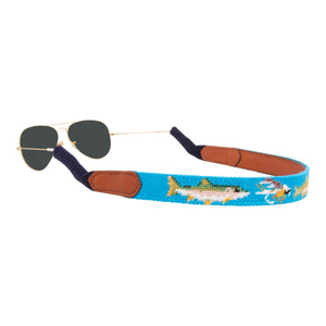 Fly Fishing Needlepoint Sunglass Strap with light blue background showing a fly-fishing themed pattern of rainbow trout, salmon, fly rods and reels, dry flies, nymph patterns, and cutthroat.  Sunglass straps have a full grain leather backing and secure cotton wrapped ear piece connectors for a secure grip. 
