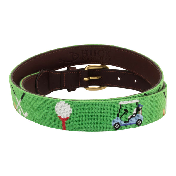 Golfing Needlepoint Belt with light green background showing a repeating pattern of teed gold ball, crossed gold clubs, and colorful golf carts. Belt has a brass buckle and full grain leather backing and strap.