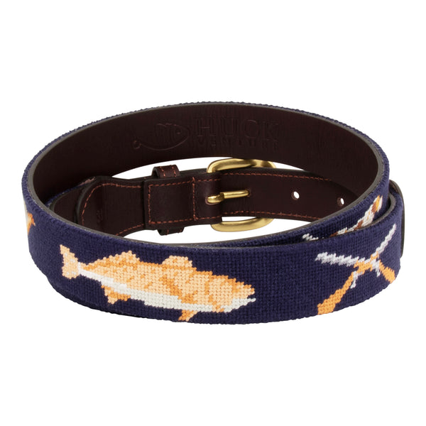 Back side of Sportsman Needlepoint Belt with navy blue background showing outdoor sporting themed images of a pointer dog, crossed shotguns, shotgun shell, fly fishing rod, redfish, fishing boat, flying duck and dove.  Belt has brass buckle and full-grain leather backing.