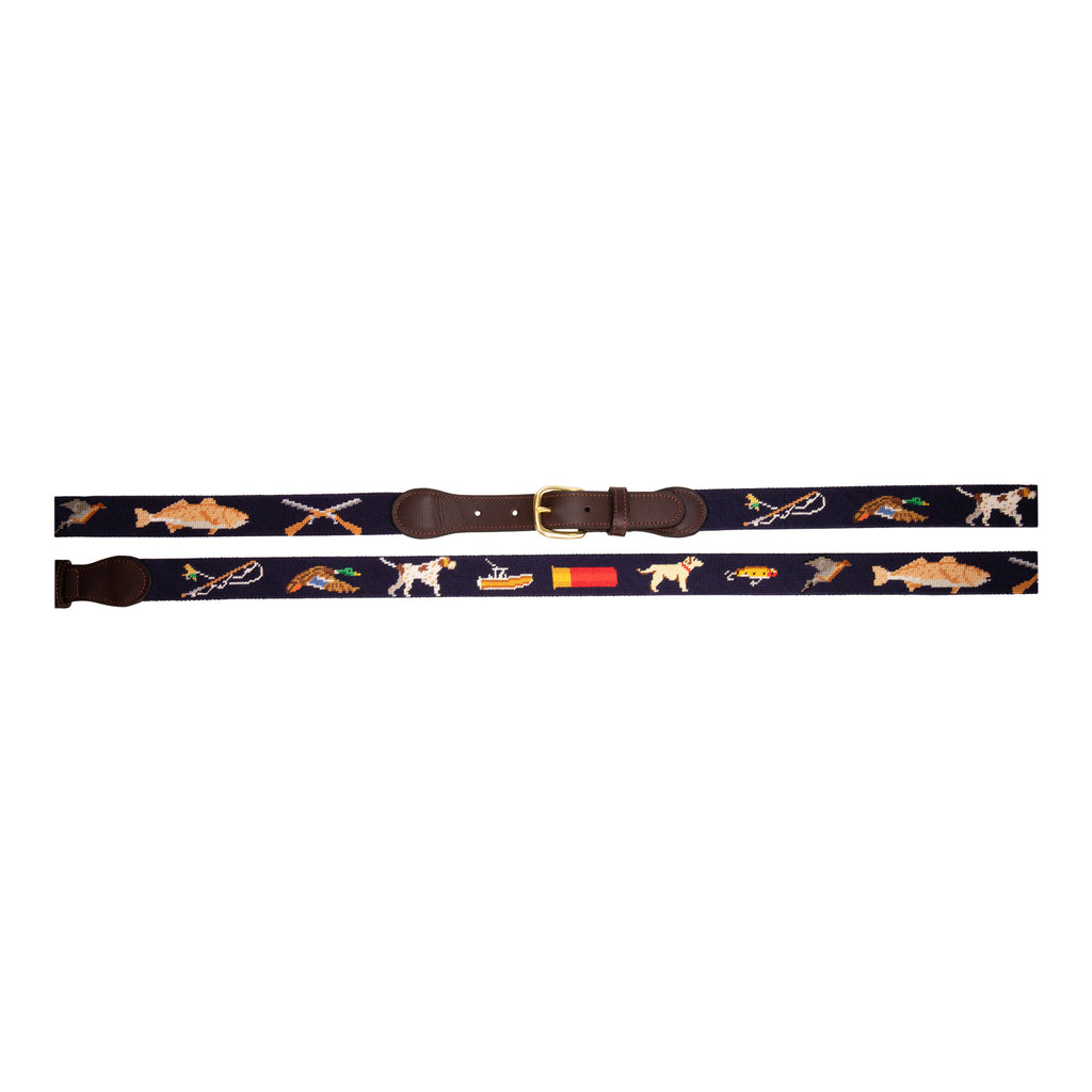 Sportsman Needlepoint Belt with navy blue background showing outdoor sporting themed images of a pointer dog, crossed shotguns, shotgun shell, fly fishing rod, redfish, fishing boat, flying duck and dove.  Belt has brass buckle and full-grain leather backing.