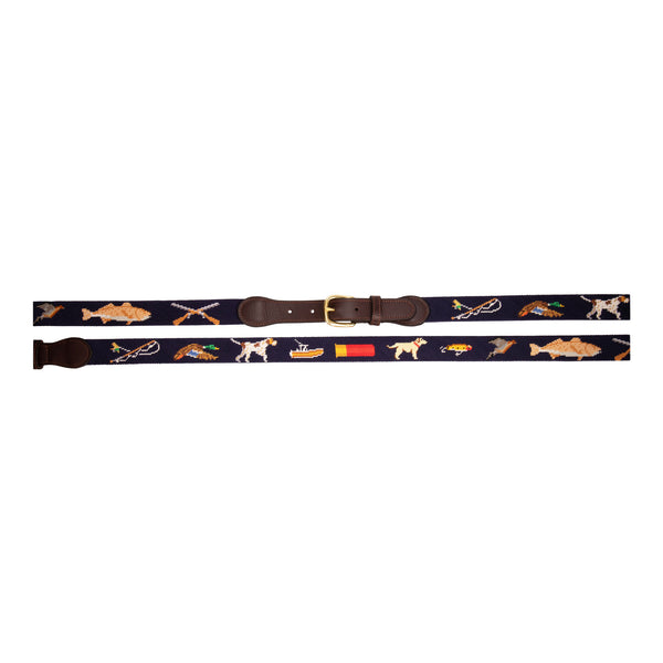 Sportsman Needlepoint Belt with navy blue background showing outdoor sporting themed images of a pointer dog, crossed shotguns, shotgun shell, fly fishing rod, redfish, fishing boat, flying duck and dove.  Belt has brass buckle and full-grain leather backing.